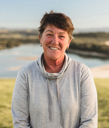 smiling woman wearing grey top, field, beach and calm water in background